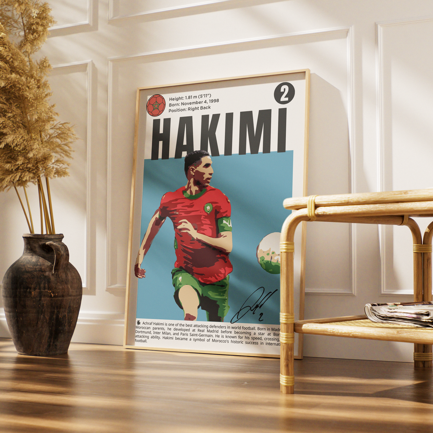 A framed vertical poster featuring a football player with the name HAKIMI in large letters, styled leaning against a white paneled wall on a wooden floor beside a rattan side table and a dark vase with dried grass.