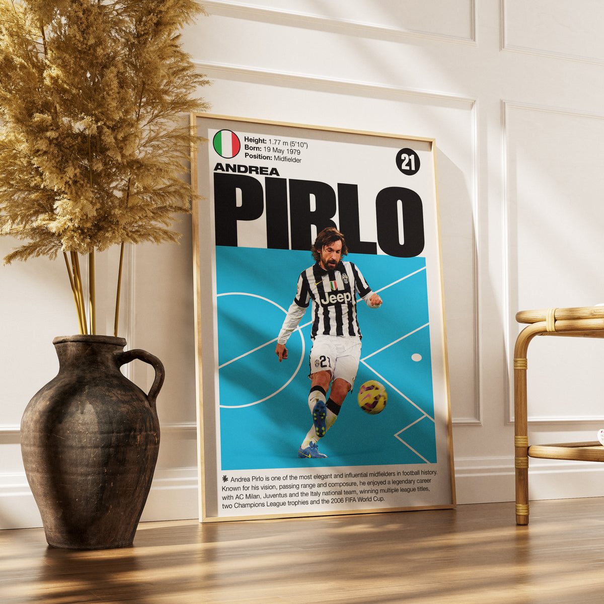A framed poster of footballer Andrea Pirlo leaning against a white paneled wall on a wooden floor, next to a dark ceramic vase with dried grass and a gold-framed chair.