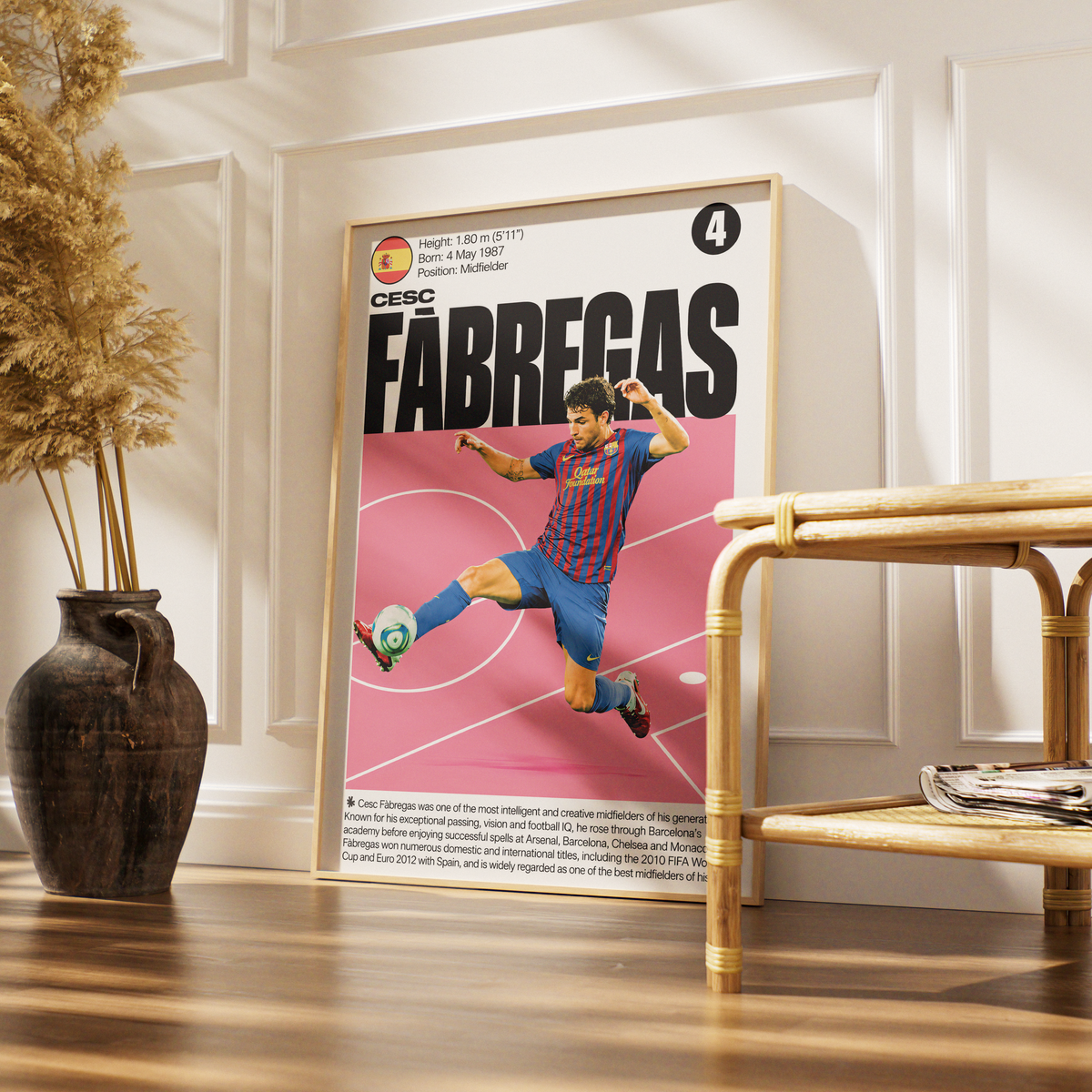 A framed vertical poster of soccer legend Lionel Messi in his FC Barcelona blue and red striped jersey, displayed on a wooden floor in a styled room next to a bamboo side table and a dark vase with dried grass.