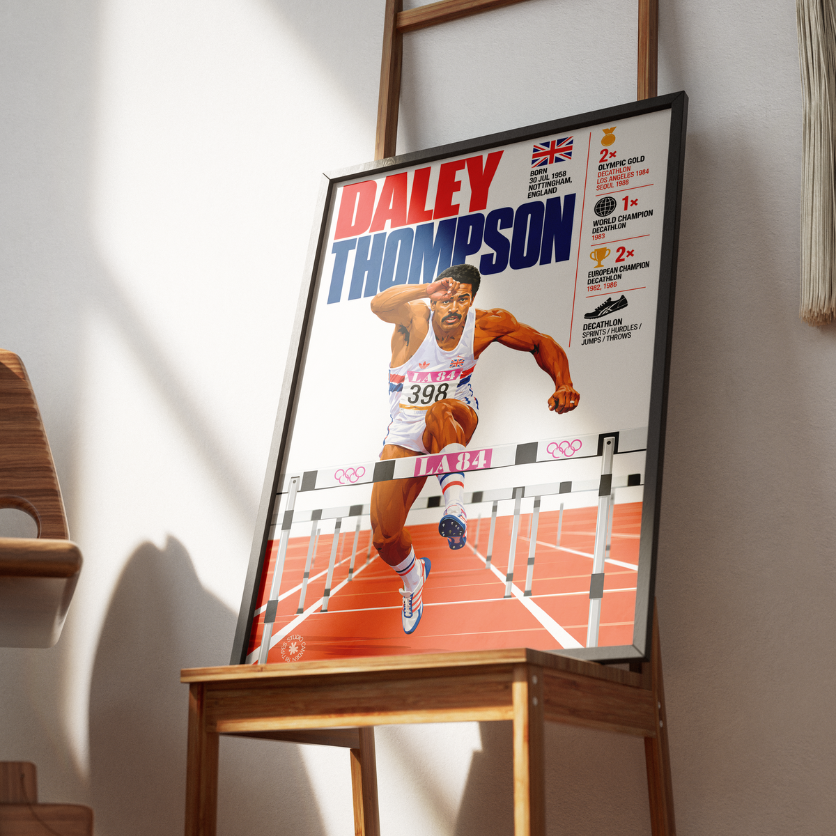 A framed poster of athlete Daley Thompson displayed on a wooden easel in a sunlit room. The poster features a colorful illustration of Thompson crossing a hurdle on a track, with his name in large red and blue text and various achievement badges on the right side.