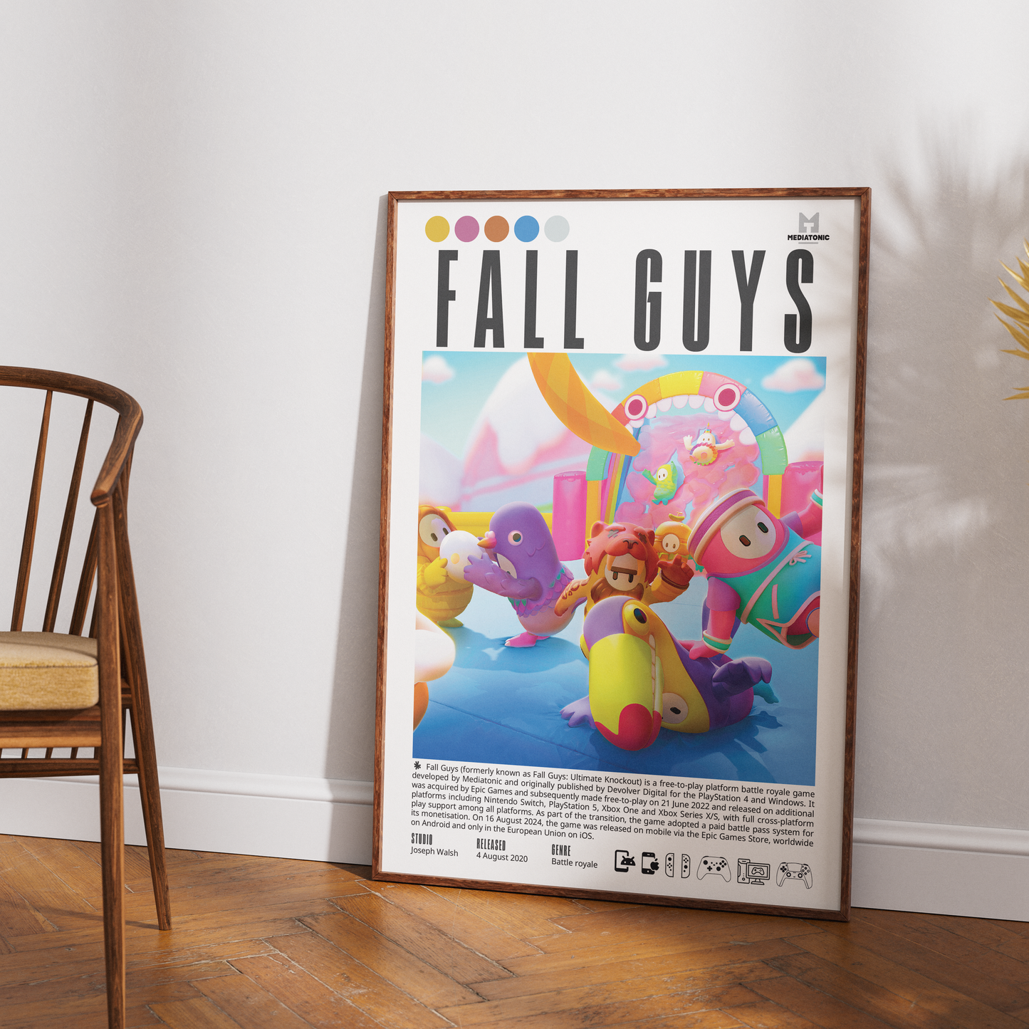 A framed vertical poster featuring the 'Fall Guys' game artwork, leaning against a white wall on a wooden floor next to a wooden chair.