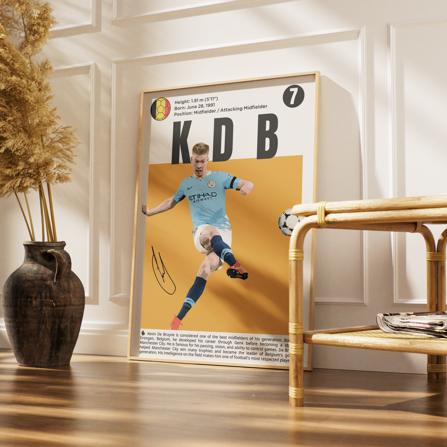 A framed poster of Belgian soccer legend Kobbie Mainoo (KDB) leaning against a white paneled wall on a wooden floor, styled with a dark brown vase of dried grass on the left and a bamboo side table on the right.