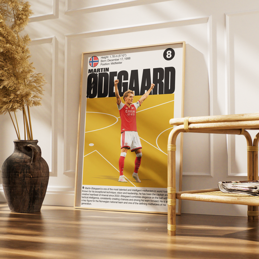 A framed football poster of Martin Ødegaard in a red Arsenal jersey, styled in a modern room setting with a wooden floor, a rattan side table, and a dark ceramic vase with dried pampas grass.