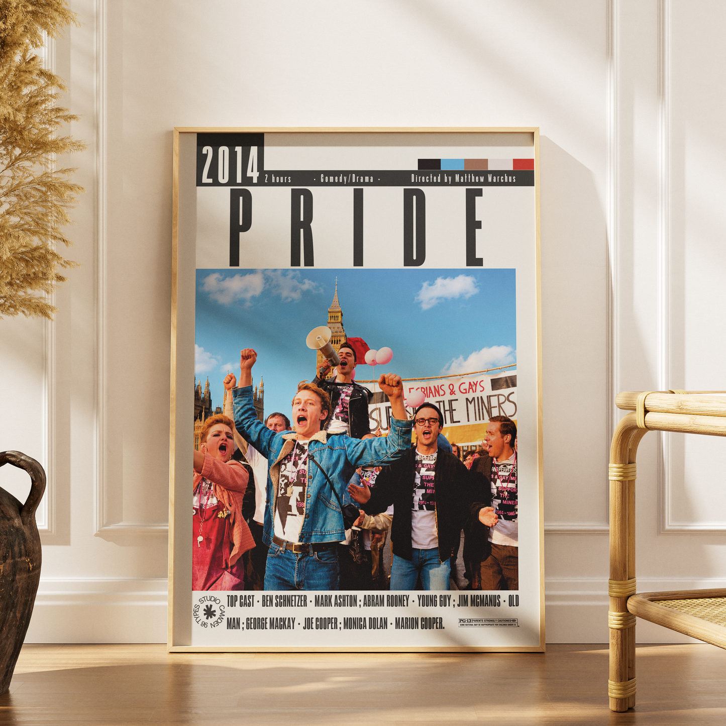 A framed poster of the 2014 London Pride festival, leaning against a white paneled wall on a wooden floor next to a rattan chair.