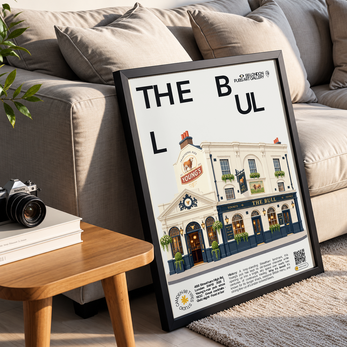 A framed poster leaning against a beige sofa in a living room setting. The poster features a black frame and a white background with the text 'THE BUL' and a stylized illustration of a pub facade labeled 'The Bul' with additional text and logos at the bottom.