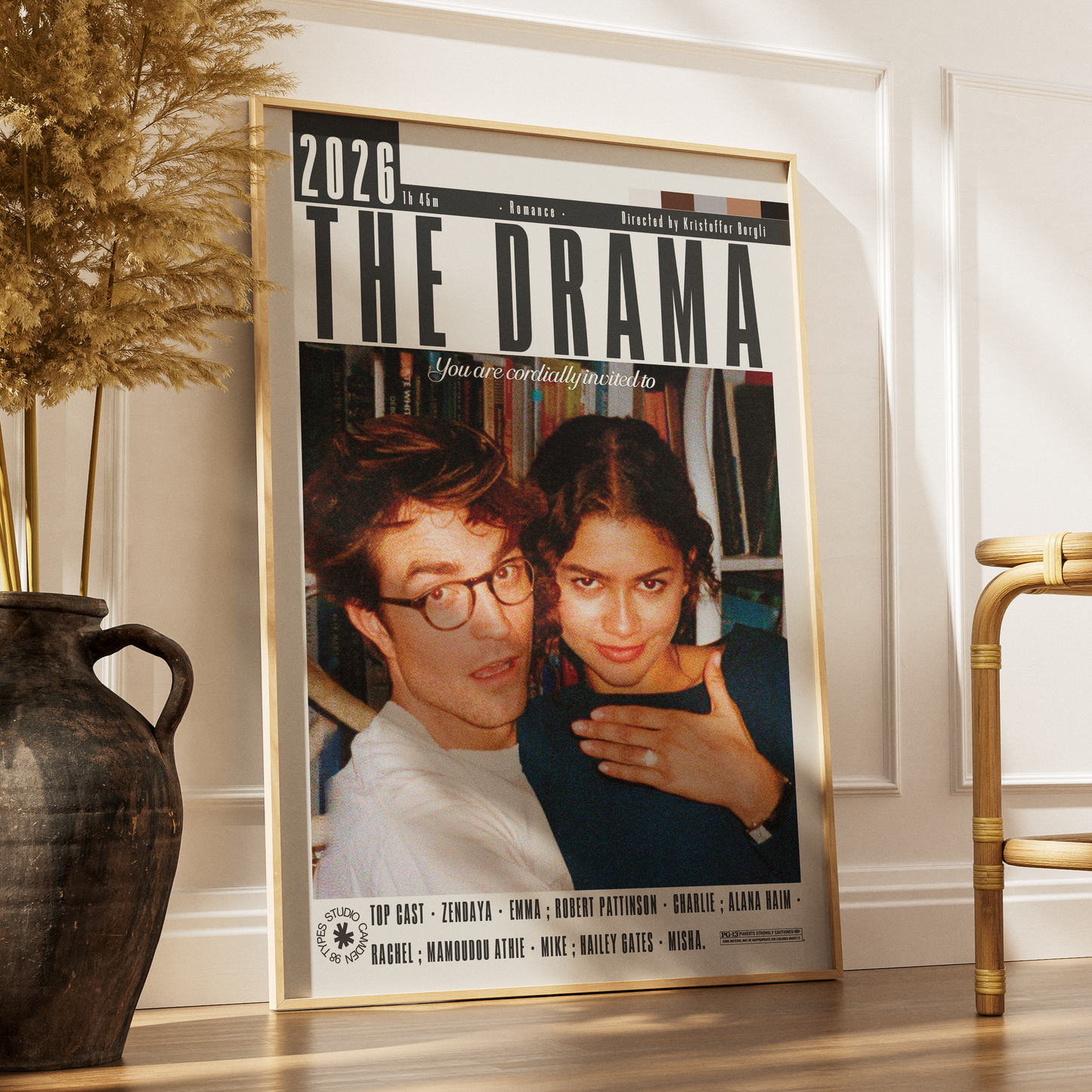 A framed vertical poster leaning against a white wall on a wooden floor, styled in a home interior next to a dark vase with dried grass and a gold chair. The poster is titled 'THE DRAMA 2026' and features a photo collage of a man and woman with movie-style text and credits.
