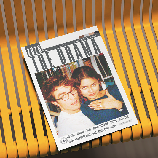 A magazine or book cover titled "THE DRAMA" lies on a yellow slatted bench. The cover features a large photo of two people and text including "2026" and "EXCITING.