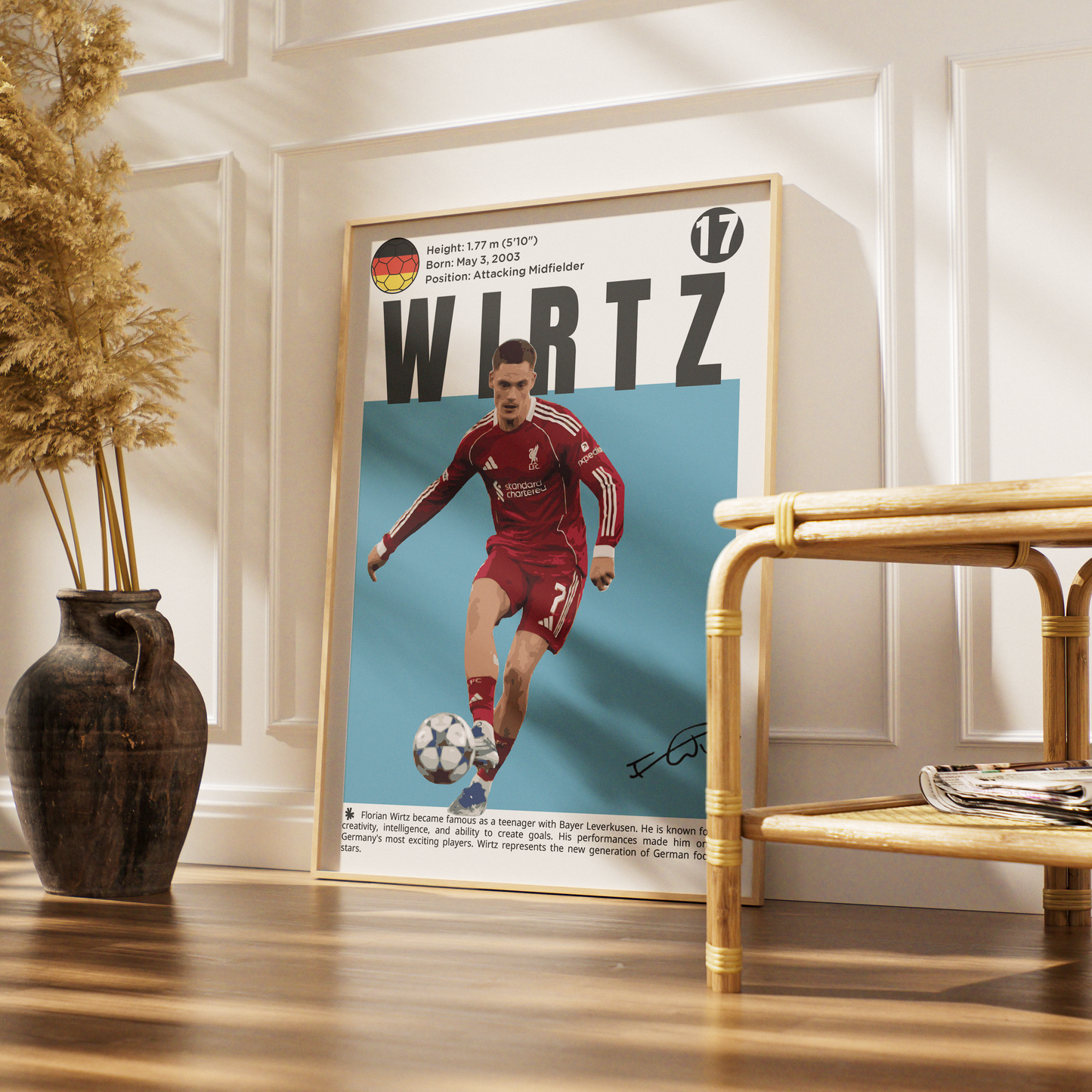 A framed vertical poster featuring a soccer player in a red Liverpool kit on a light blue background, leaning against a white paneled wall in a styled interior with a wooden floor, vase, and rattan furniture.