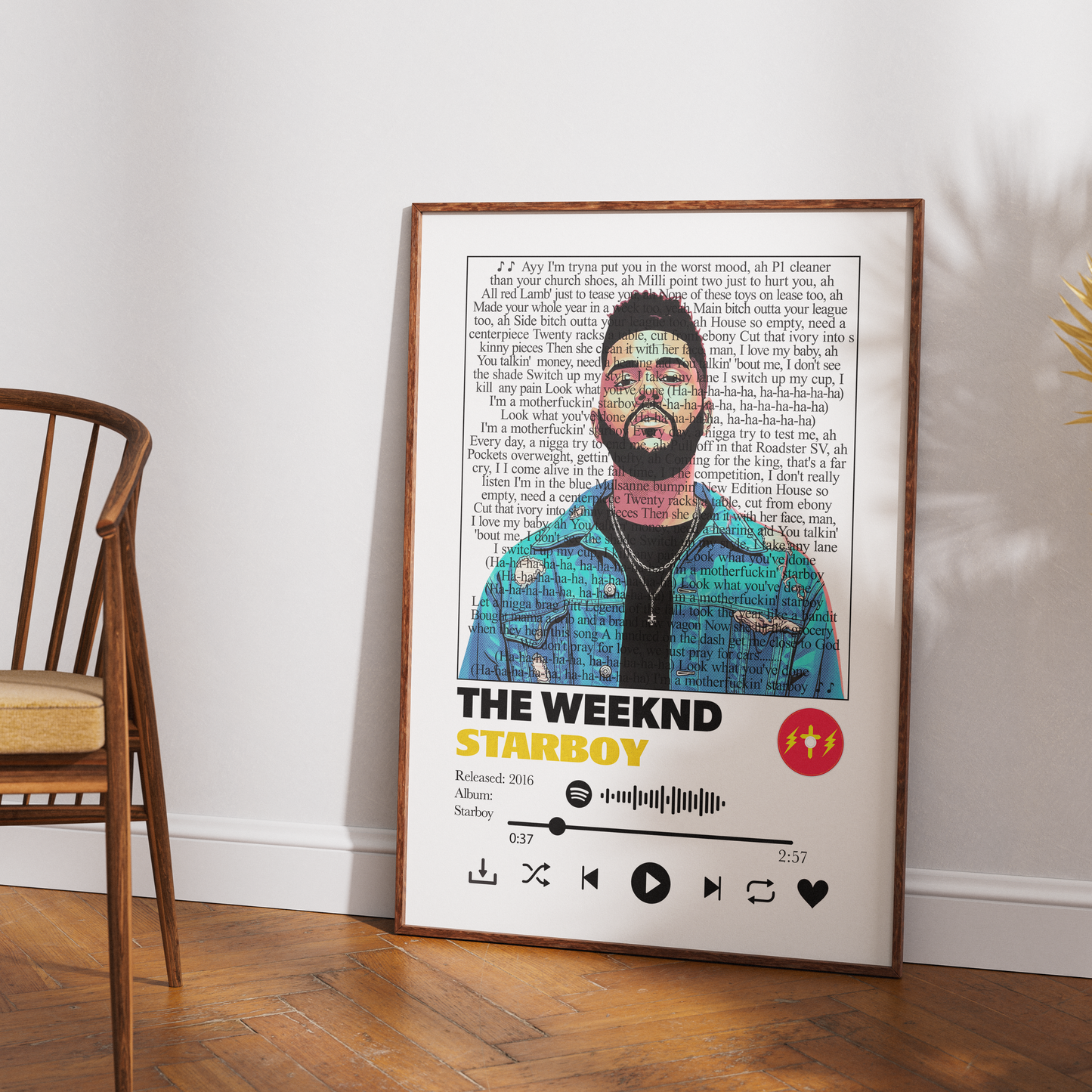 A framed music poster of The Weeknd's 'Starboy' leaning against a white wall on a wooden floor, next to a wooden chair.