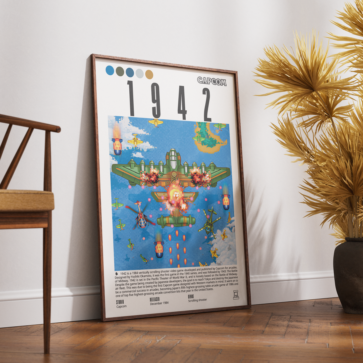 A framed vintage-style poster leaning against a white wall on a wooden floor, next to a chair and a potted plant. The poster features the title “1942” and a colorful illustration from Ghibli’s Spirited Living with a CAPCOM logo at the top right.