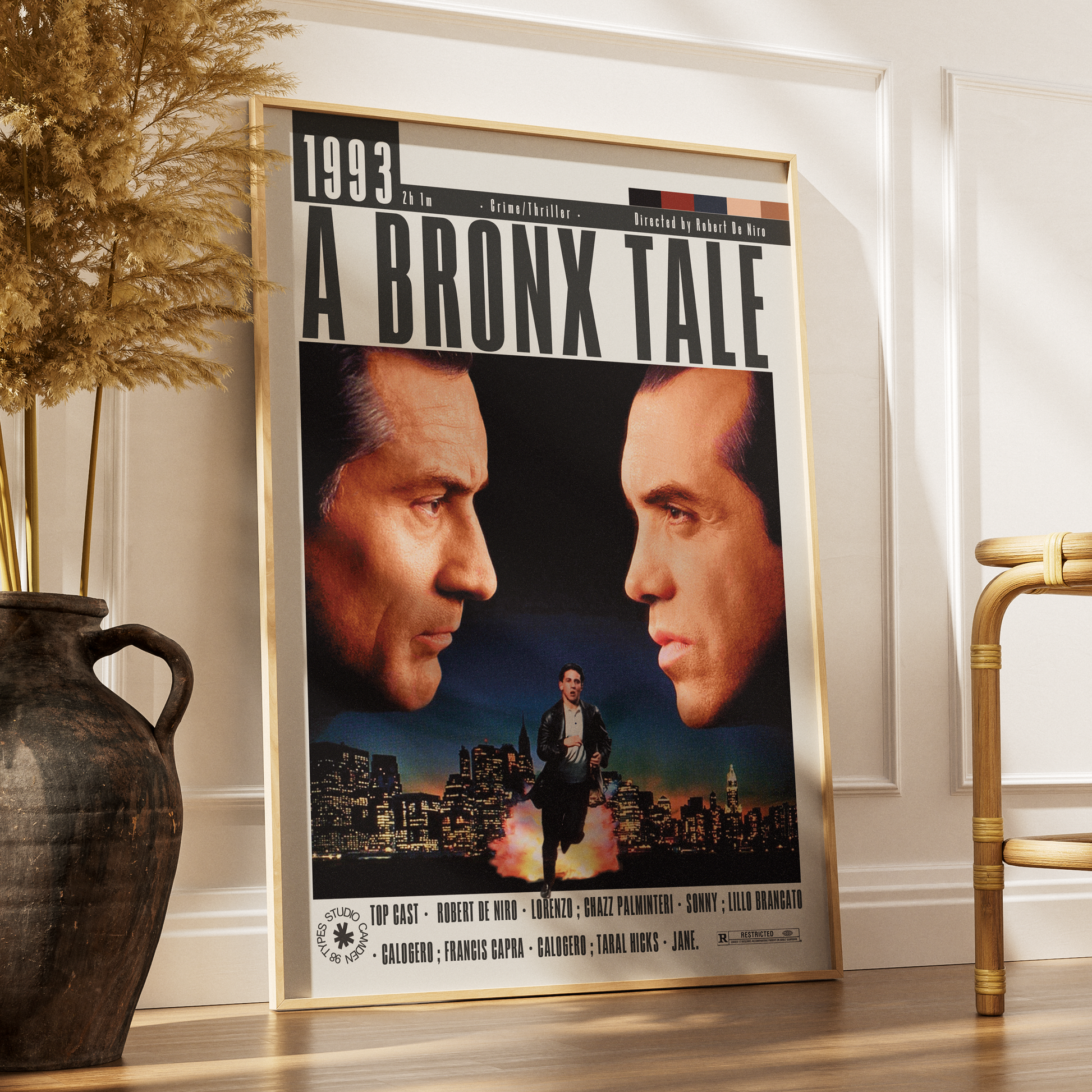 A framed movie poster for 'A Bronx Tale' leaning against a white wall on a wooden floor, styled in a home interior next to a dark ceramic vase with dried grass and a gold chair.