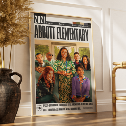 A framed 2021 Abbott Elementary movie poster leaning against a white wall on a wooden floor, next to a dark vase with dried grass and part of a gold chair.
