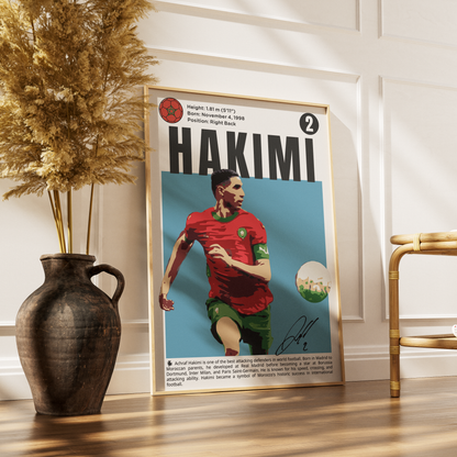 A framed poster featuring a stylized portrait of footballer Hakimi in a red Portugal jersey, leaning against a white paneled wall on a wooden floor beside a dark vase with dried grass and a gold chair.