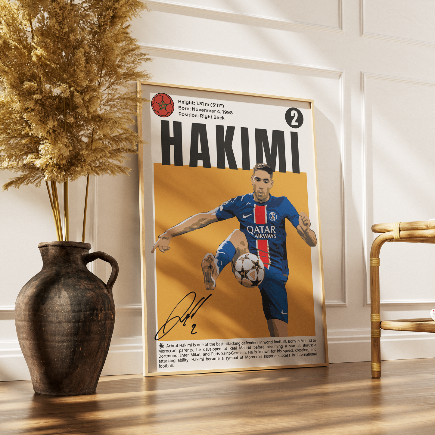 A framed soccer poster featuring a portrait of Patrick Hakimi in a Paris Saint-Germain jersey, leaning against a white paneled wall on a wooden floor next to a dark ceramic vase with dried grass and a gold chair.