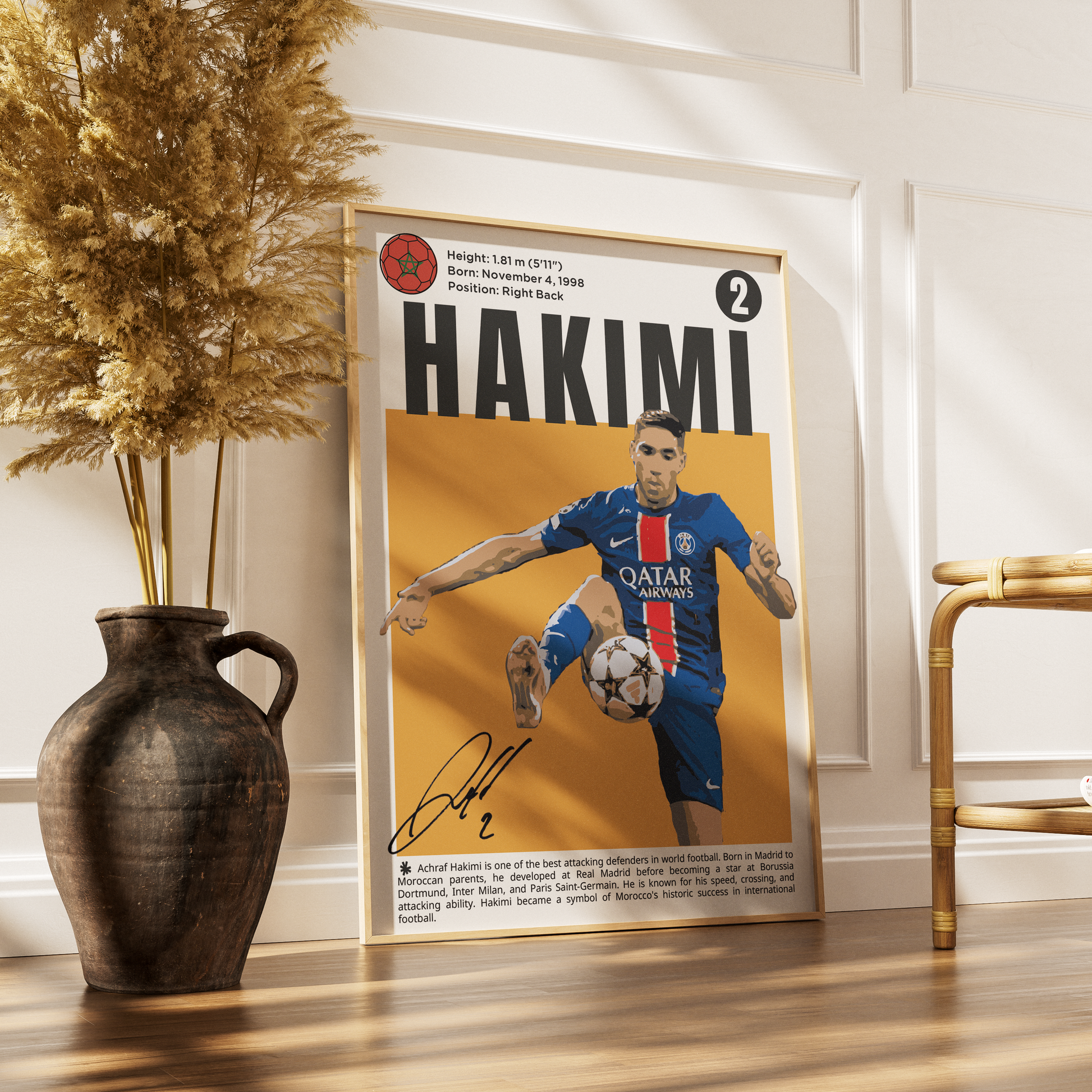 A framed soccer poster featuring a portrait of Patrick Hakimi in a Paris Saint-Germain jersey, leaning against a white paneled wall on a wooden floor next to a dark ceramic vase with dried grass and a gold chair.