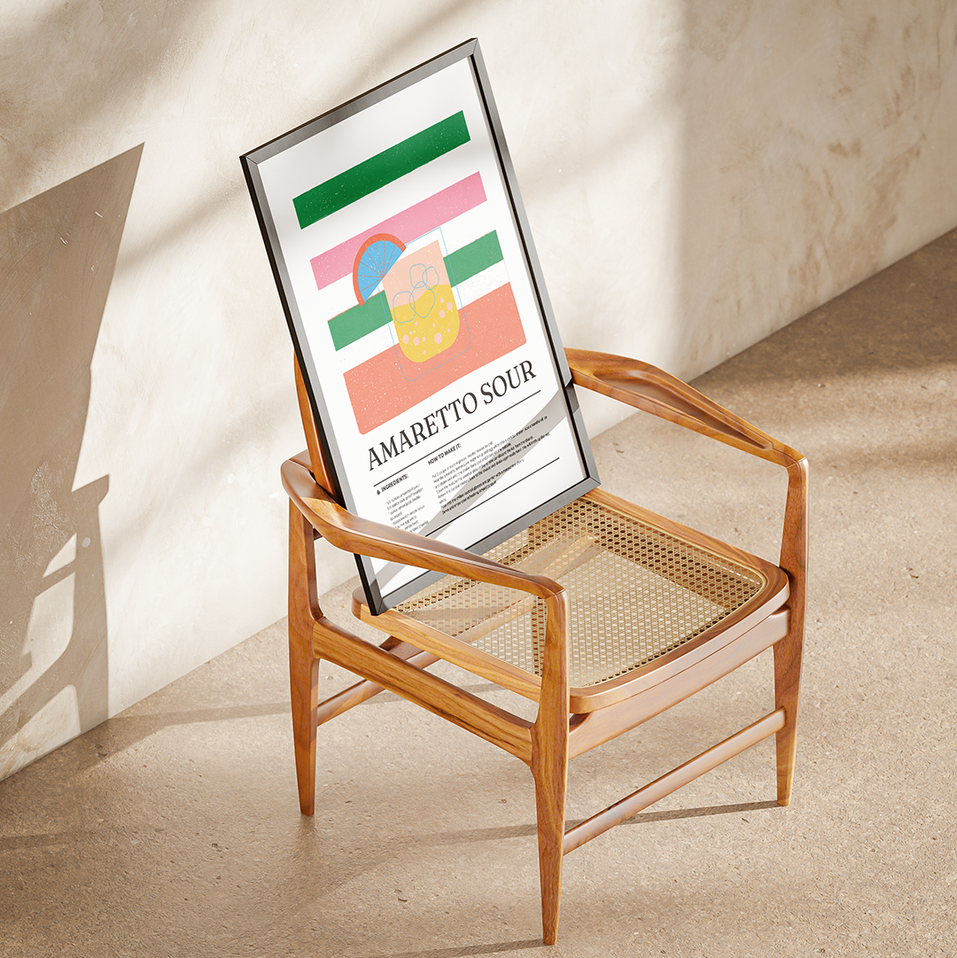 A framed art print with a colorful abstract design and the text 'AMARETTO SOUR' leaning on a mid-century modern wooden armchair in a sunlit room.