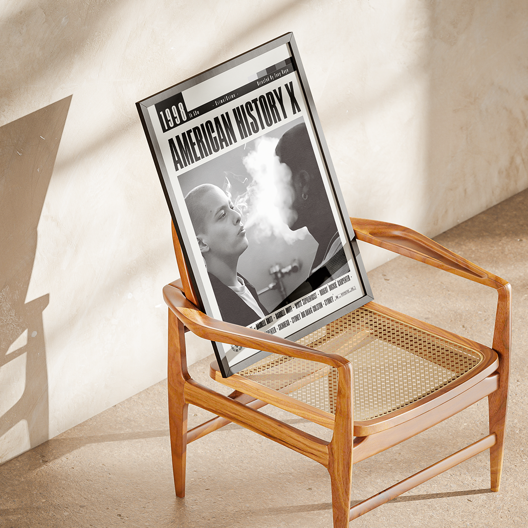 A framed black-and-white poster titled 'American History X' leaning against a wooden armchair in a sunlit room with a textured beige wall and floor.