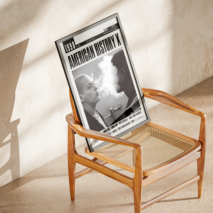A framed black-and-white poster titled 'American History X' leaning against a wooden armchair in a sunlit room with a textured beige wall and floor.
