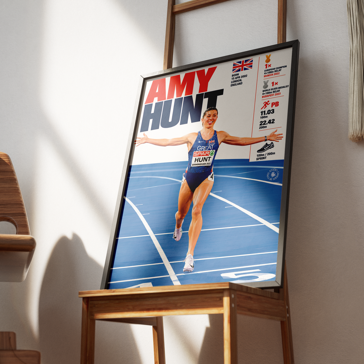A framed poster featuring athlete Amy Hunt on a blue running track, displayed on a wooden stand in a real indoor setting with sunlight and shadows.