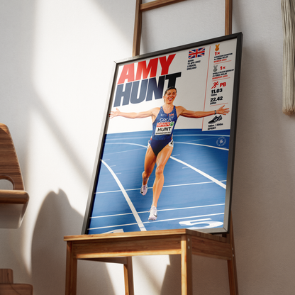 A framed poster featuring athlete Amy Hunt on a blue running track, displayed on a wooden stand in a real indoor setting with sunlight and shadows.