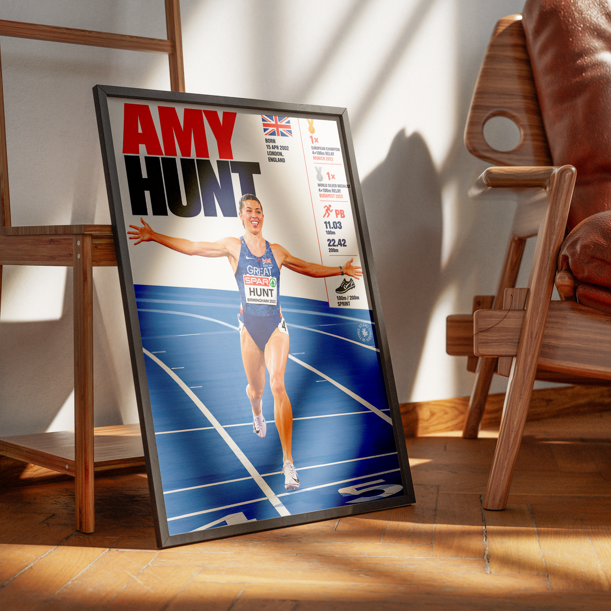 A framed poster of British athlete Amy Hunt leaning against a wall on a wooden floor in a modern room setting with wooden chairs.