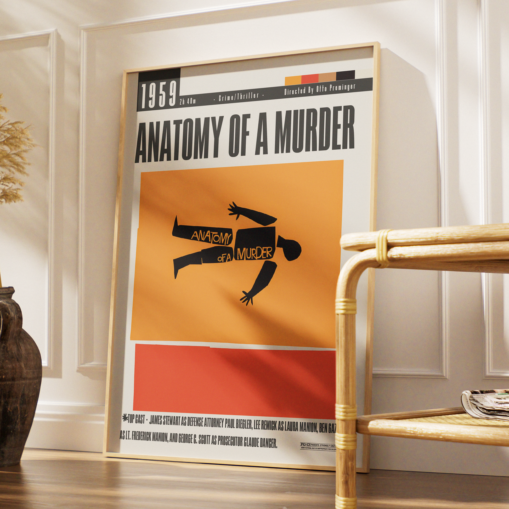 A framed poster of the 1959 film 'Anatomy of a Murder' leaning against a white paneled wall on a wooden floor, next to a rattan side table.