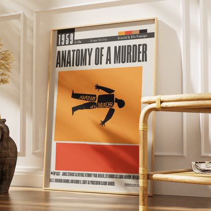 A framed poster of the 1959 film 'Anatomy of a Murder' leaning against a white paneled wall on a wooden floor, next to a rattan side table.