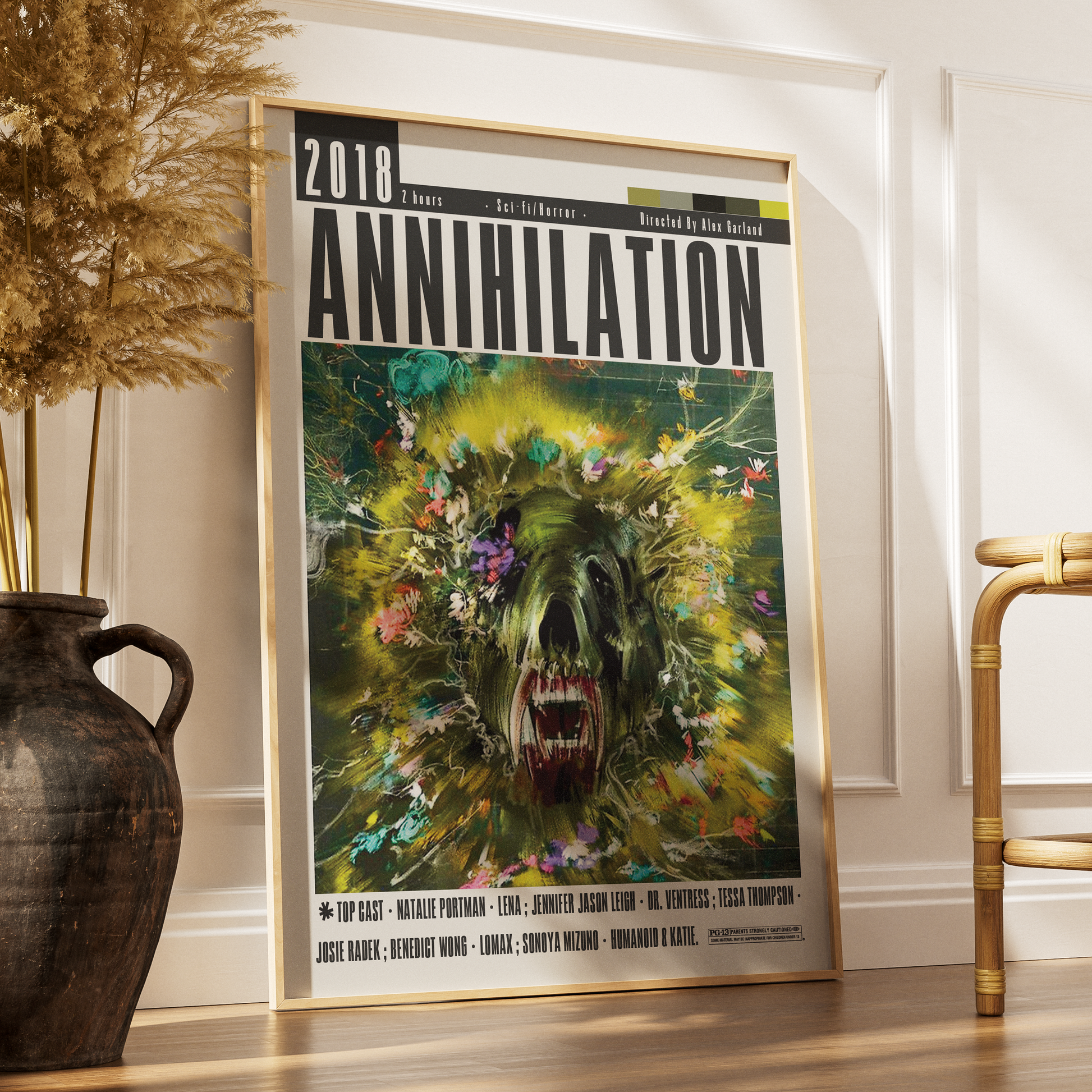 A framed 2018 Annihilation movie poster leaning against a white wall on a wooden floor, styled in a room with a dark vase of dried grass on the left and a wooden chair on the right.