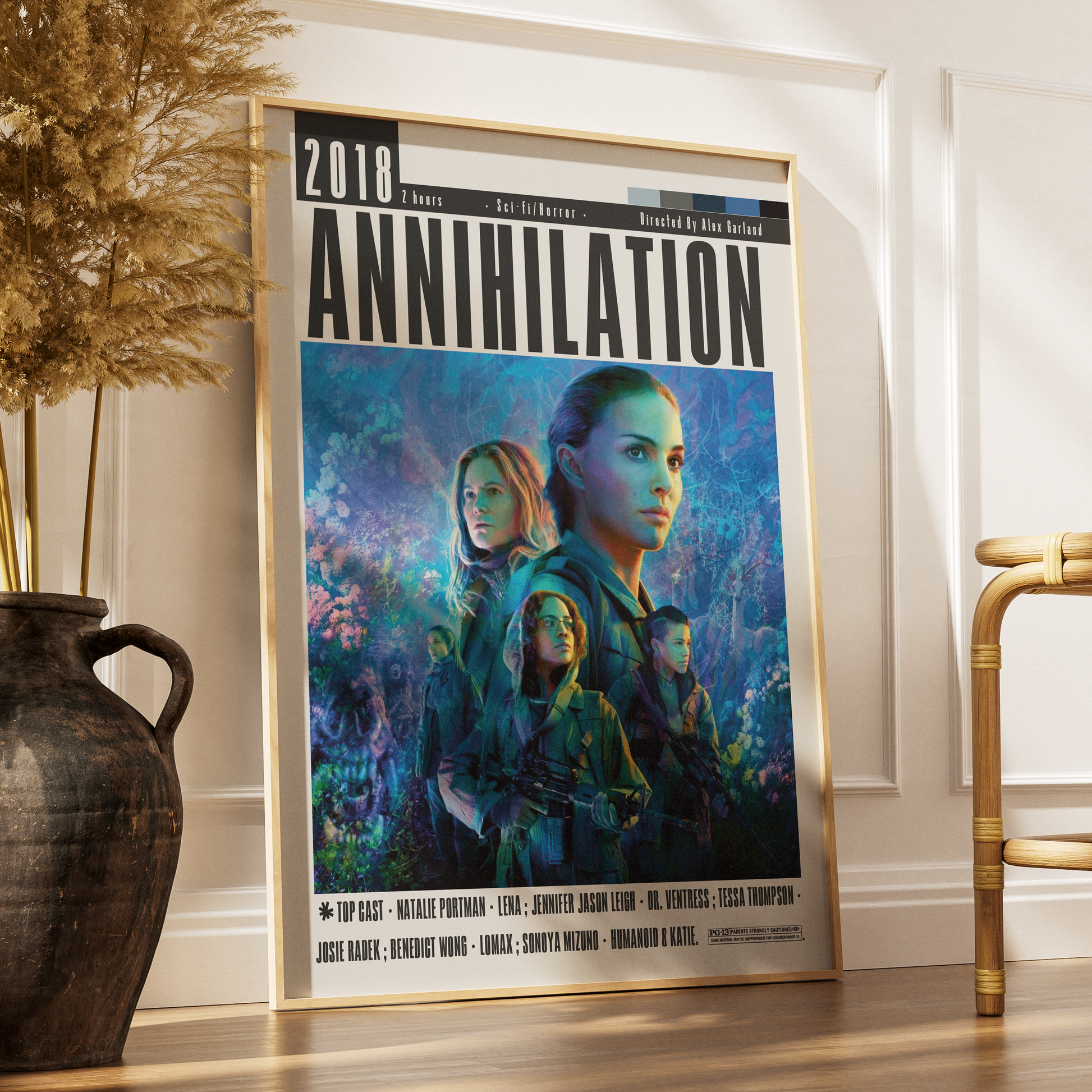 A framed movie poster for the 2018 film 'Annihilation' leaning against a white wall on a wooden floor, styled in a room setting with a dark vase of dried grass on the left and a gold chair on the right.