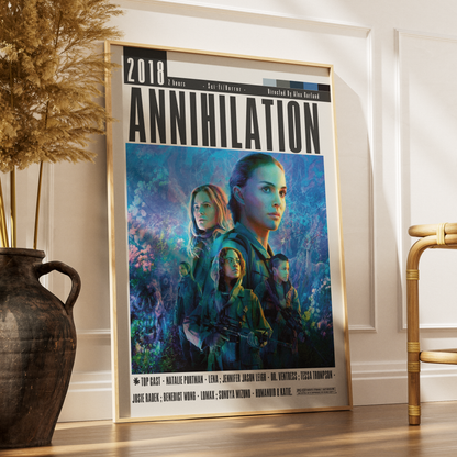A framed movie poster for the 2018 film 'Annihilation' leaning against a white wall on a wooden floor, styled in a room setting with a dark vase of dried grass on the left and a gold chair on the right.