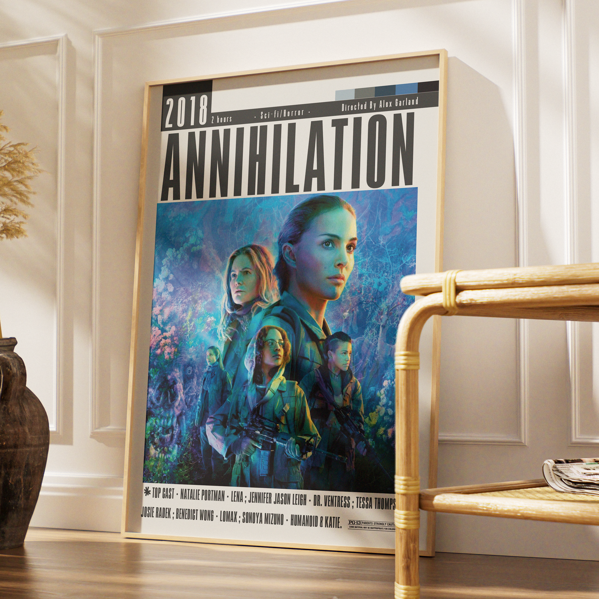 A framed movie poster for the 2018 film 'Annihilation' leaning against a white paneled wall on a wooden floor, next to a bamboo side table.