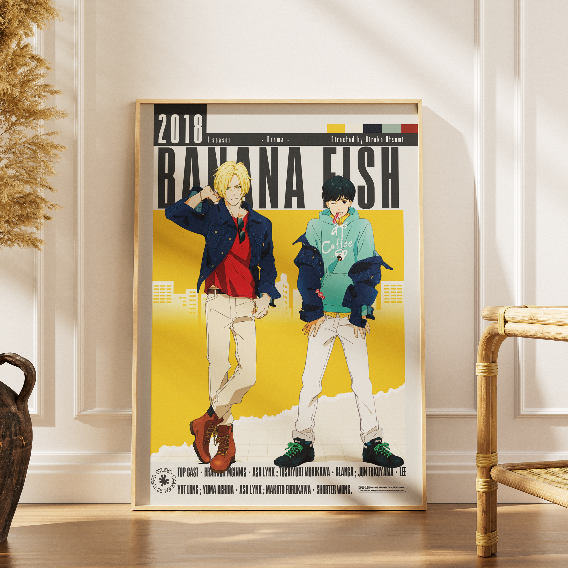 A framed anime poster of the movie '2018 Banana Fish' leaning against a white paneled wall on a wooden floor, with a dried plant and a bamboo chair visible in the room.