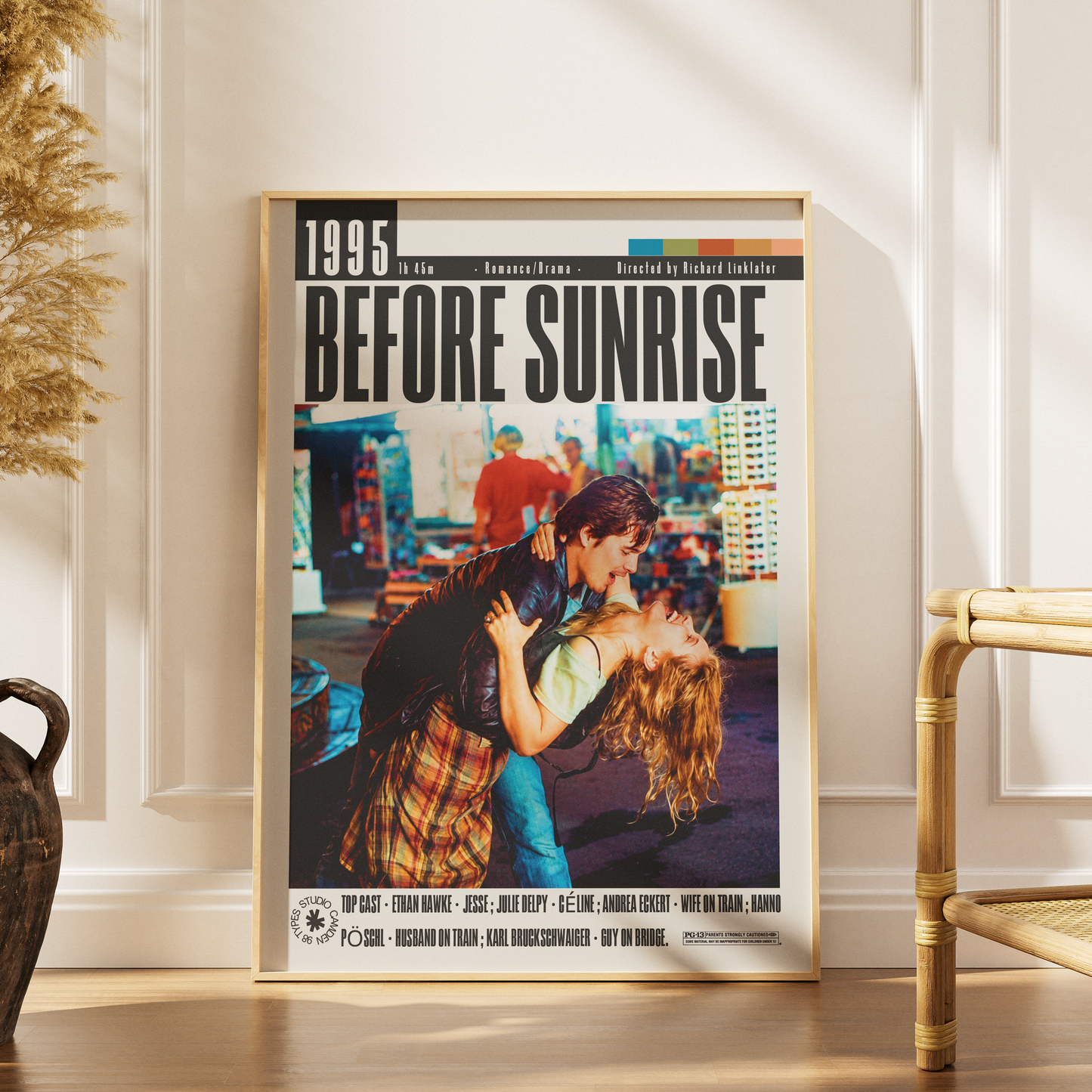 A framed movie poster for 'Before Sunrise' leaning against a white paneled wall on a wooden floor, with a woven chair and dried plant visible in the room.