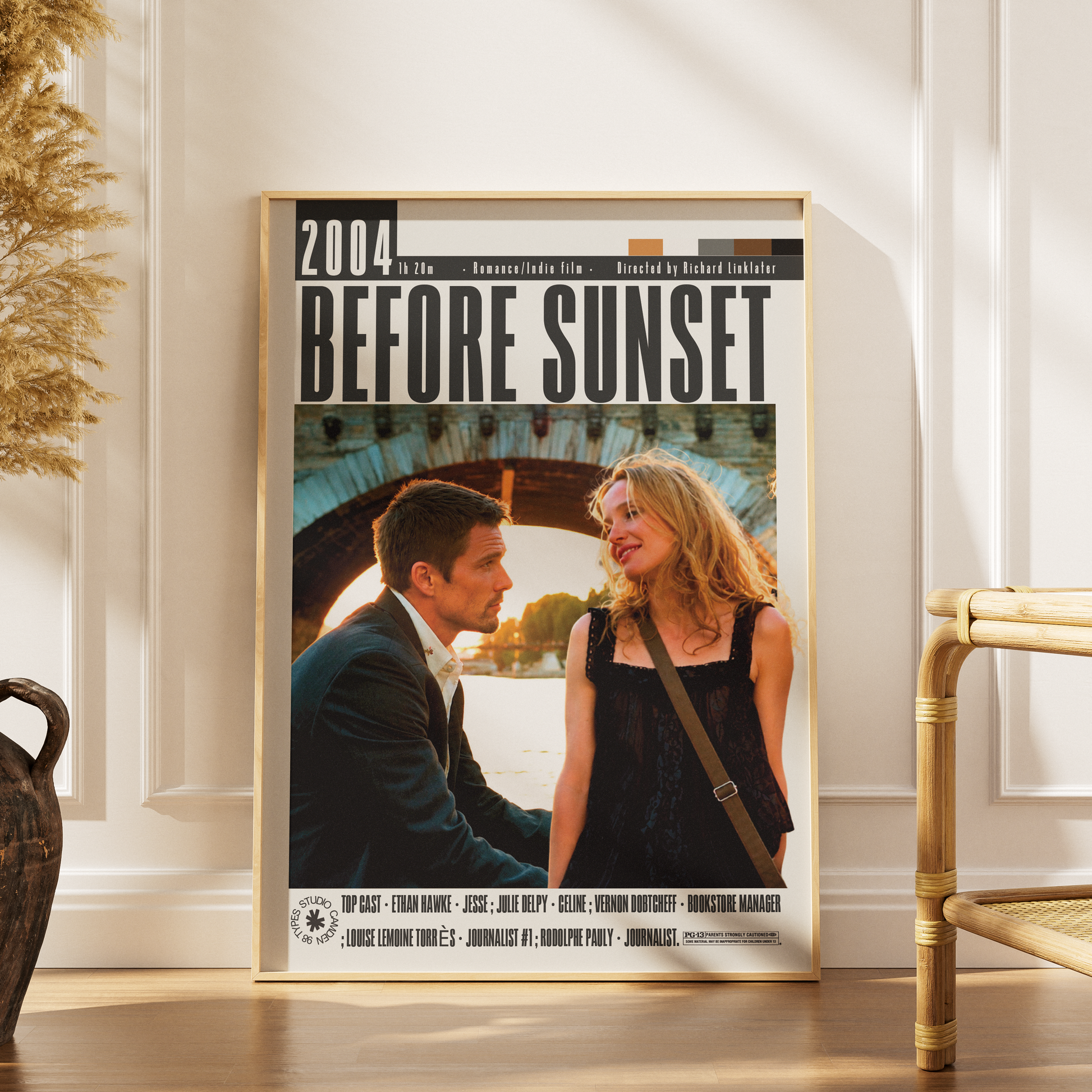 A framed movie poster for the 2004 film 'Before Sunset' leaning against a white paneled wall on a wooden floor, with a woven chair and dried plant visible in the room.
