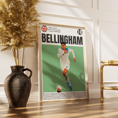 A framed football poster featuring Bellingham in a white kit against a green background, leaning against a white paneled wall on a wooden floor next to a dark vase with dried grass and a gold chair.