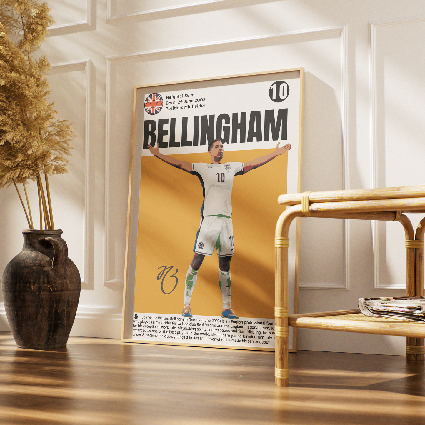 A framed football poster of Bellingham, number 10, displayed in a styled interior setting with a wooden floor, a decorative vase with dried grass, and a rattan side table.