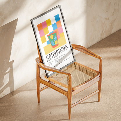 A mid-century modern wooden armchair with a woven cane seat and backrest, styled in a sunlit room with a framed poster leaning against the backrest. The poster features a colorful geometric pattern and the text 'CAIPIRINHA'.
