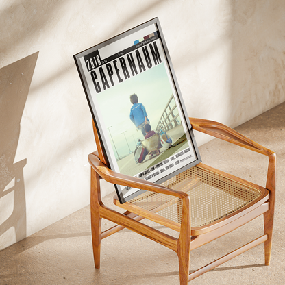 A framed poster featuring the book cover of 'Capernaum' by 2000, displayed leaning against a mid-century modern wooden armchair in a sunlit room.