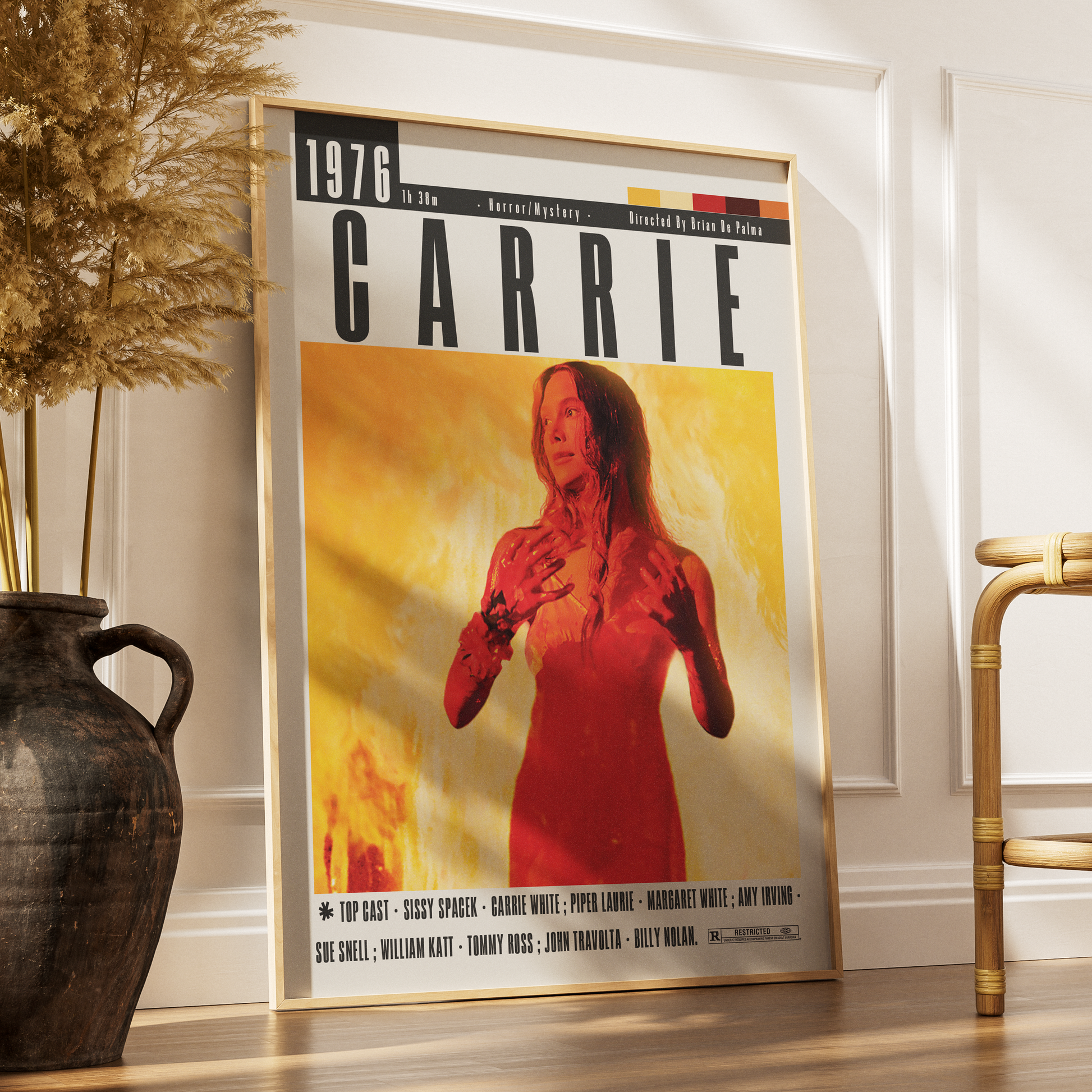 A framed poster of the movie Carrie leaning against a white wall on a wooden floor, styled in a room with a dark ceramic vase on the left and a bamboo chair on the right.