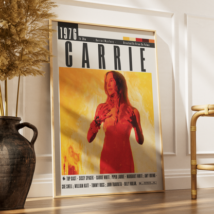 A framed poster of the movie Carrie leaning against a white wall on a wooden floor, styled in a room with a dark ceramic vase on the left and a bamboo chair on the right.