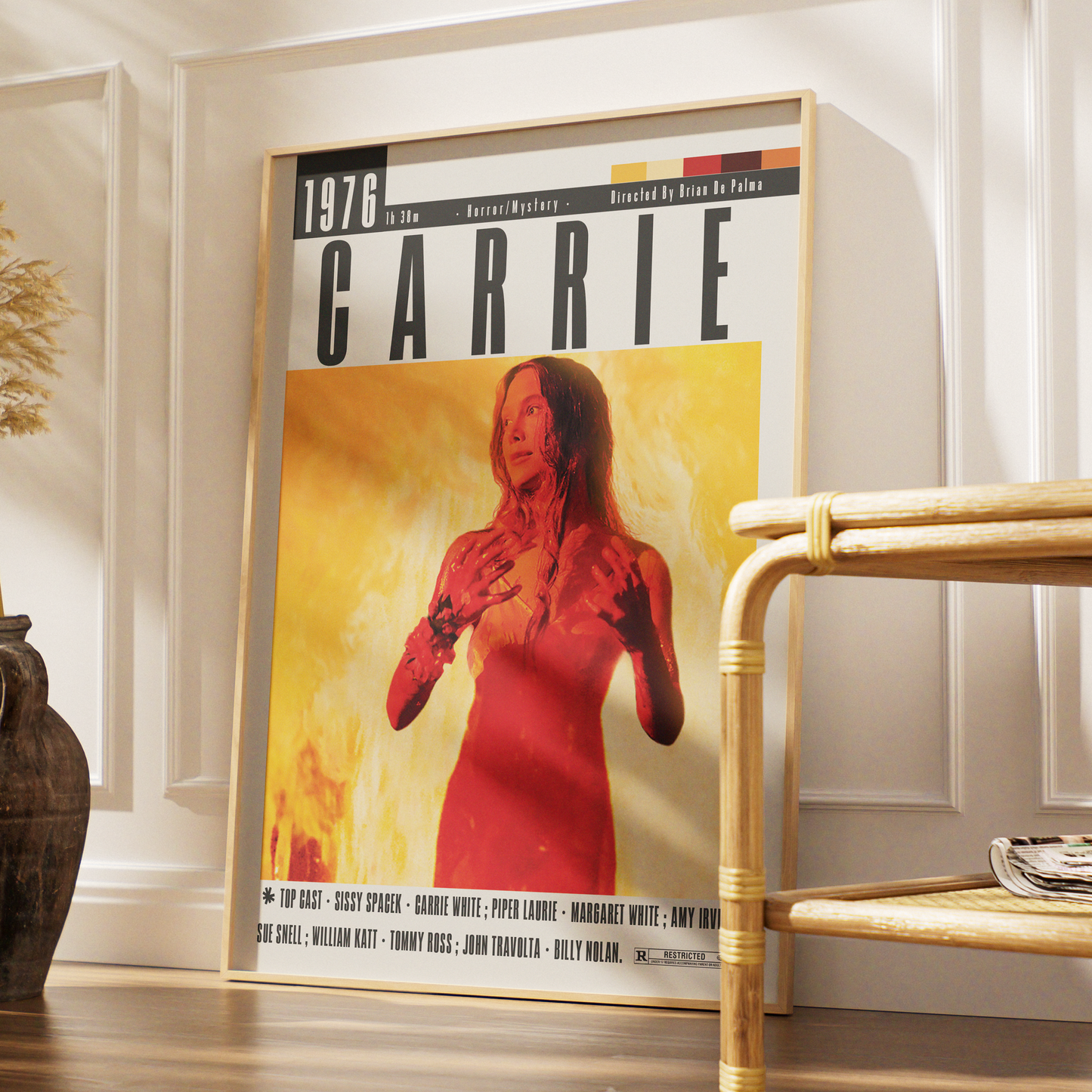 A framed poster of the movie Carrie leaning against a white paneled wall in a styled interior setting, with a wooden chair and a dark vase nearby.