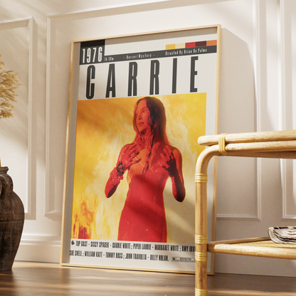 A framed poster of the movie Carrie leaning against a white paneled wall in a styled interior setting, with a wooden chair and a dark vase nearby.