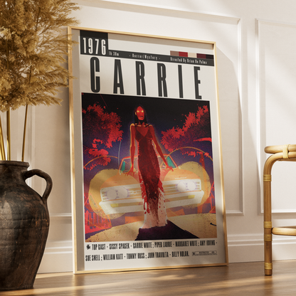 A framed poster of the classic film Carrie, styled on a wooden floor against a white paneled wall next to a dark ceramic vase with dried grass and a gold chair.