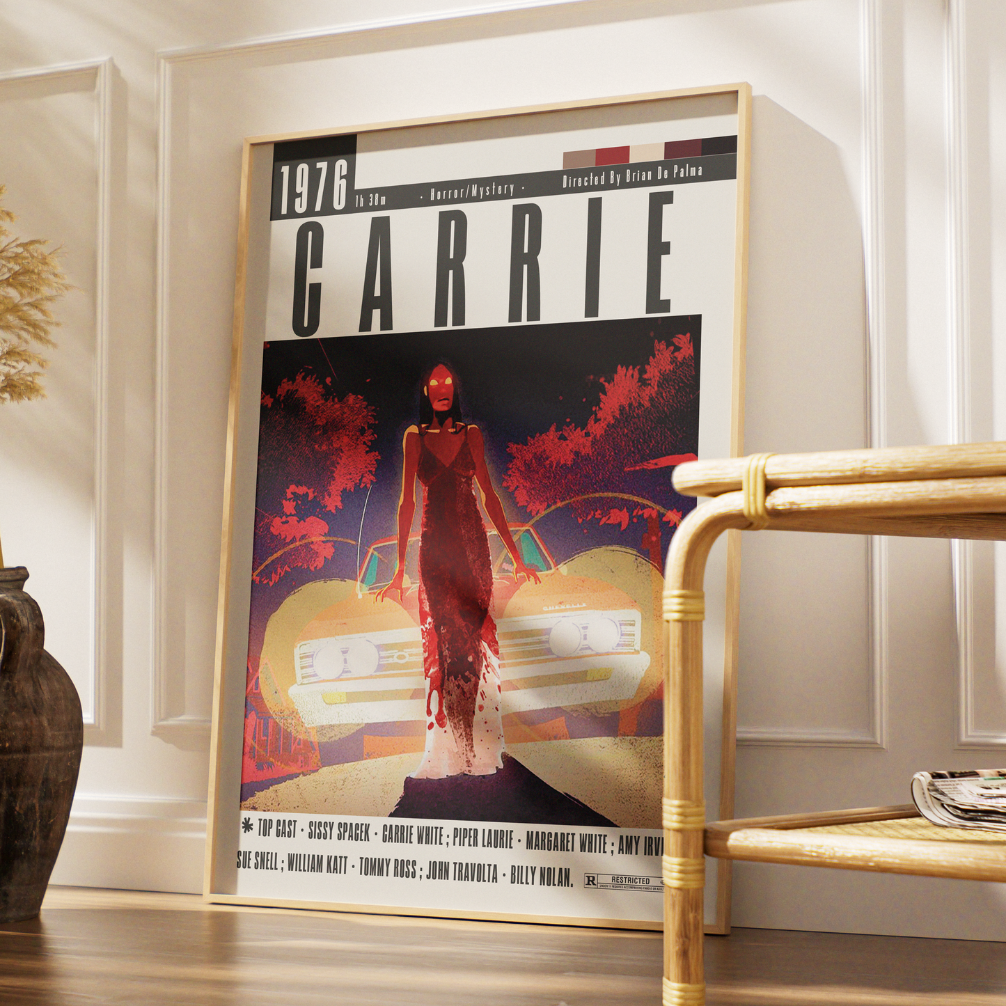 A framed poster of the movie Carrie leaning against a white paneled wall on a wooden floor, with a woven chair partially visible on the right.