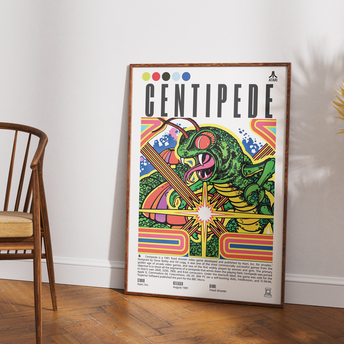 A framed Centipede poster leaning against a white wall on a wooden floor, next to a wooden chair. The poster features a colorful retro illustration of a green centipede with pink eyes and orange accents, with the title 'CENTIPEDE' in bold black letters at the top.