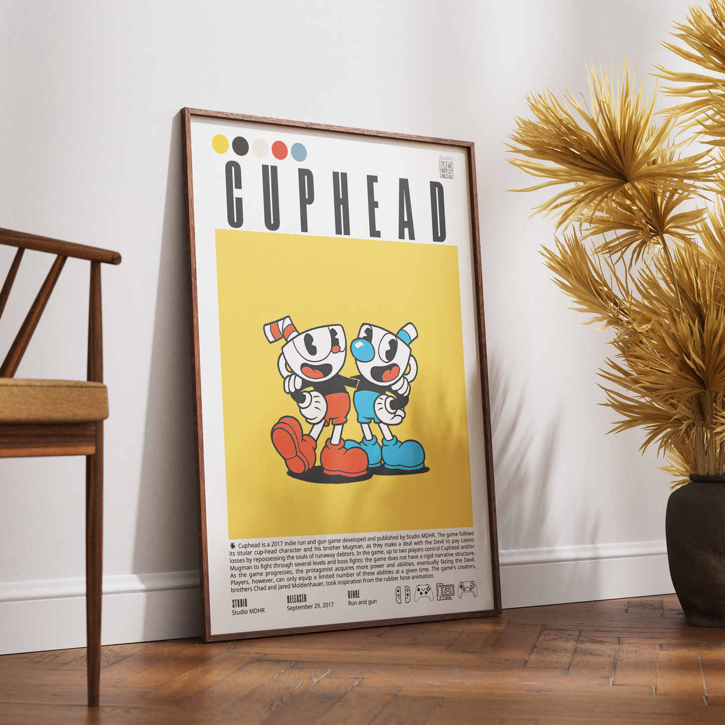 A framed Cuphead poster leaning against a white wall on a wooden floor, with a wooden chair on the left and a potted palm on the right.