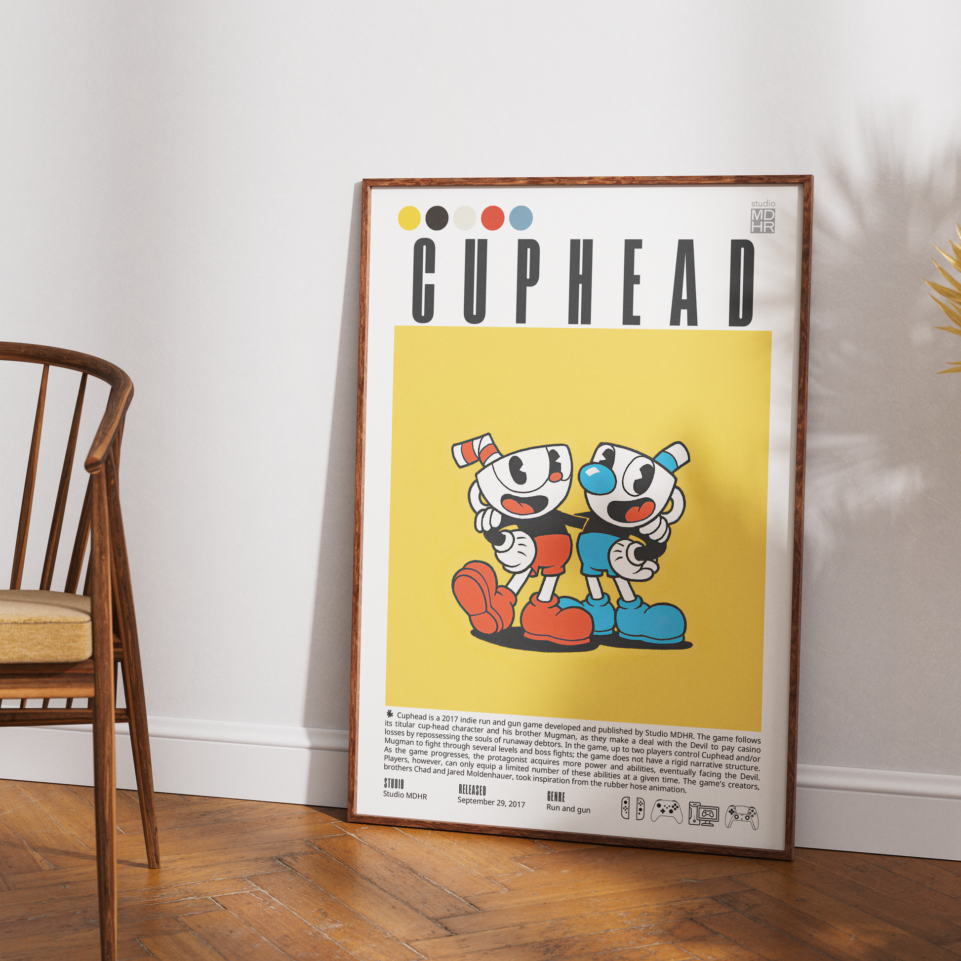 A framed CUPHEAD poster leaning against a white wall on a wooden floor, with part of a wooden chair visible on the left. The poster features the CUPHEAD title at the top and two cartoon characters on a yellow background.