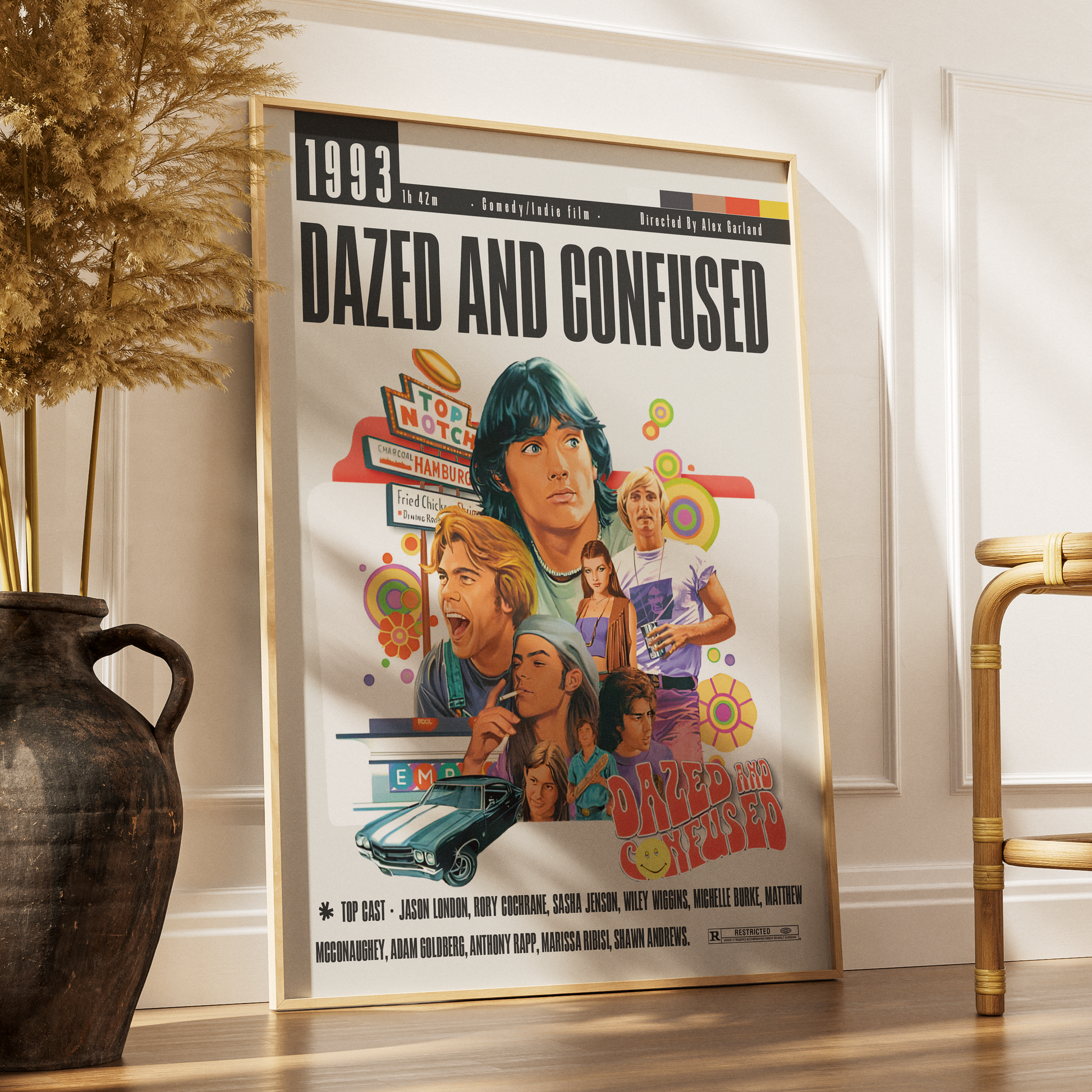 A framed 'Dazed and Confused' movie poster leaning against a white wall on a wooden floor, styled in a room setting with a dark ceramic vase and dried grass on the left and a gold-framed chair on the right.