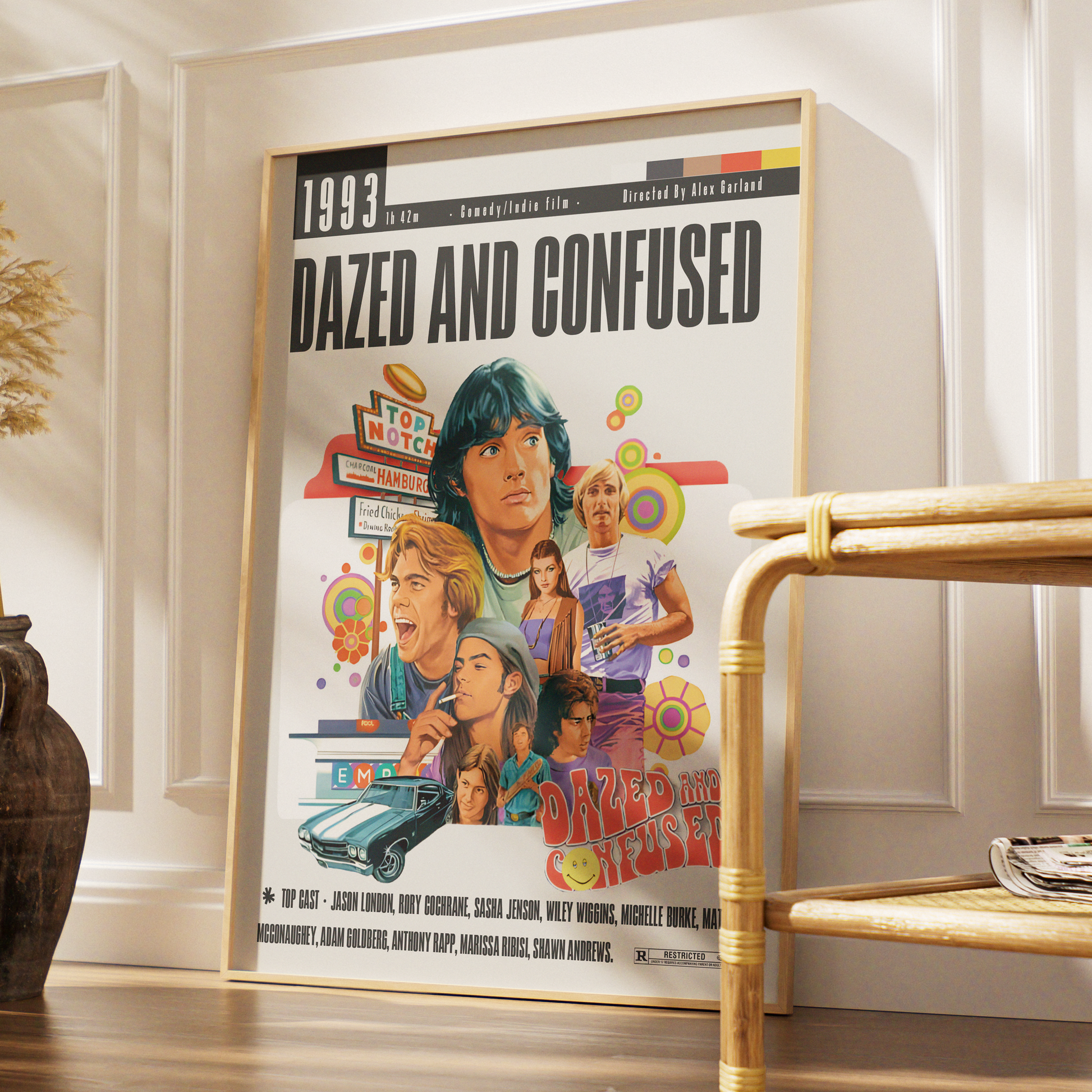 A framed 'Dazed and Confused' movie poster leaning against a white paneled wall in a styled interior setting with a bamboo side table and wooden floor.