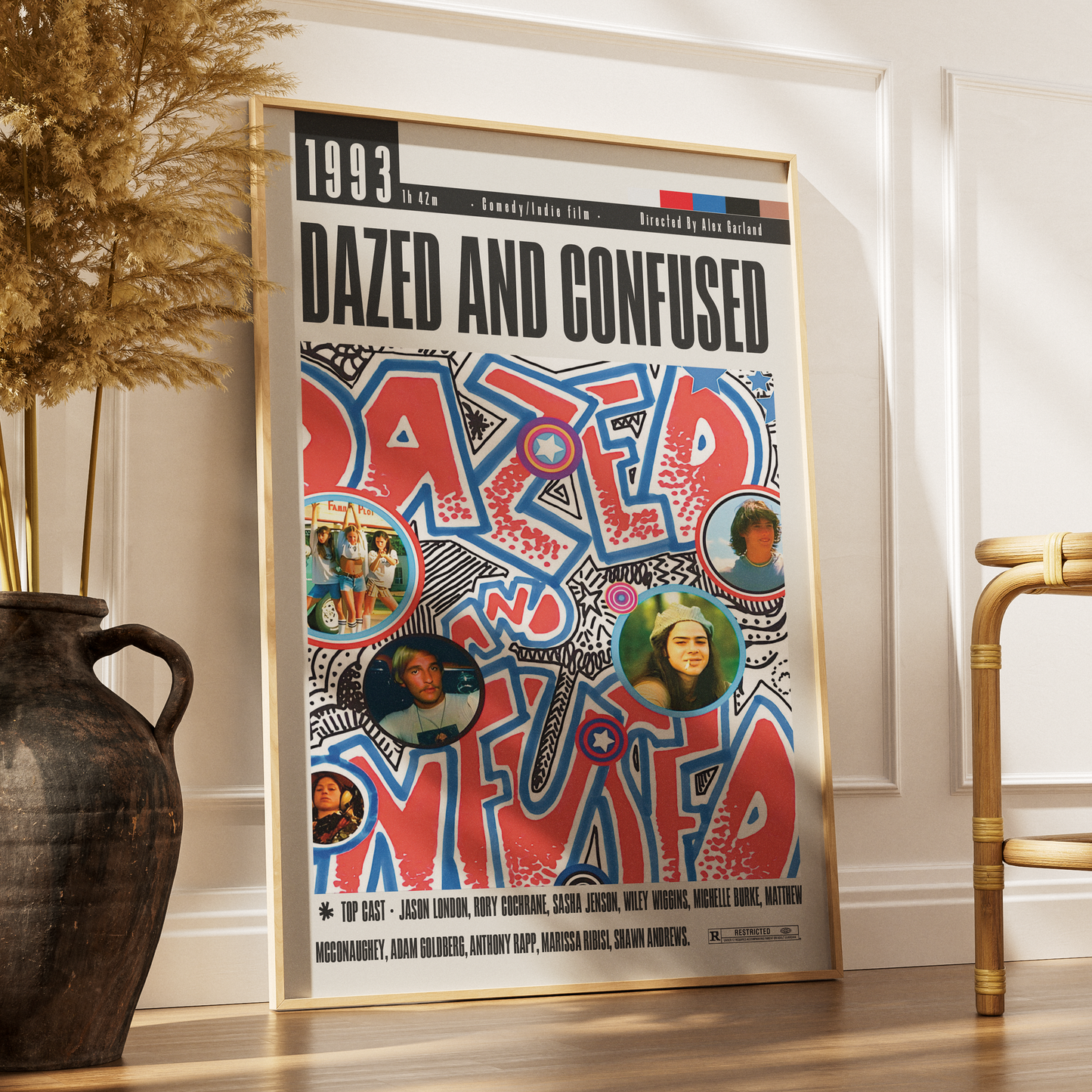 A framed music poster for the band 'Dazed and Confused' from 1993, leaning against a white wall on a wooden floor next to a dark vase with dried grass and part of a woven chair.